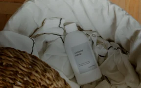 Unbranded laundry detergent bottle in a basket with white linens on a wooden floor.